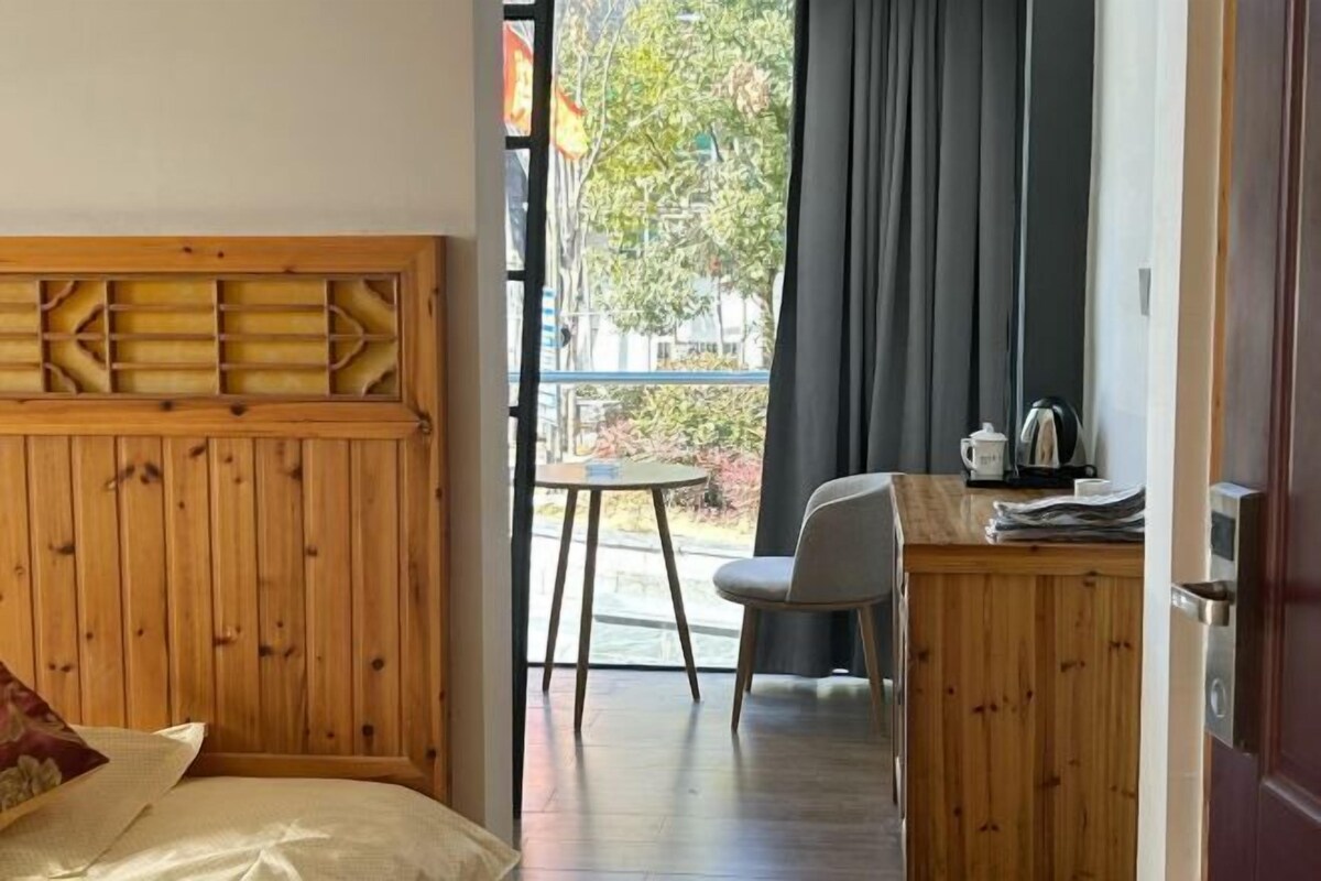 Una habitación con cama, cabecera de madera, una mesita con silla, cocina y vistas al exterior.