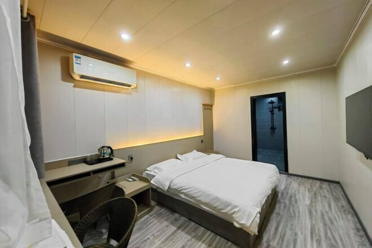 A hotel room with a bed, bedside table, chair, desk, and an air conditioning unit.
