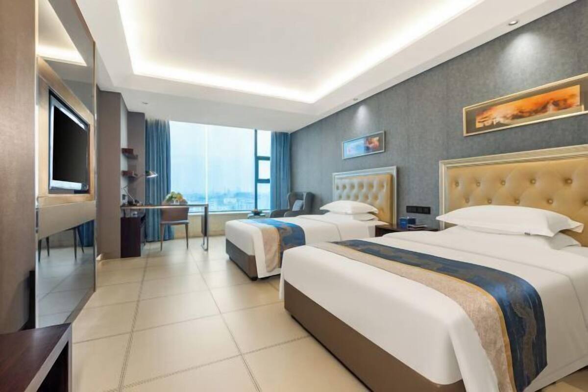 A hotel room with two beds, a desk, a chair, a TV, and a large window offering a city view.