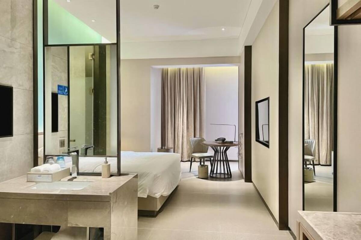 A modern hotel room with a large bed, a bathroom with a vanity, a dining area with a table and chairs, and a large mirror.