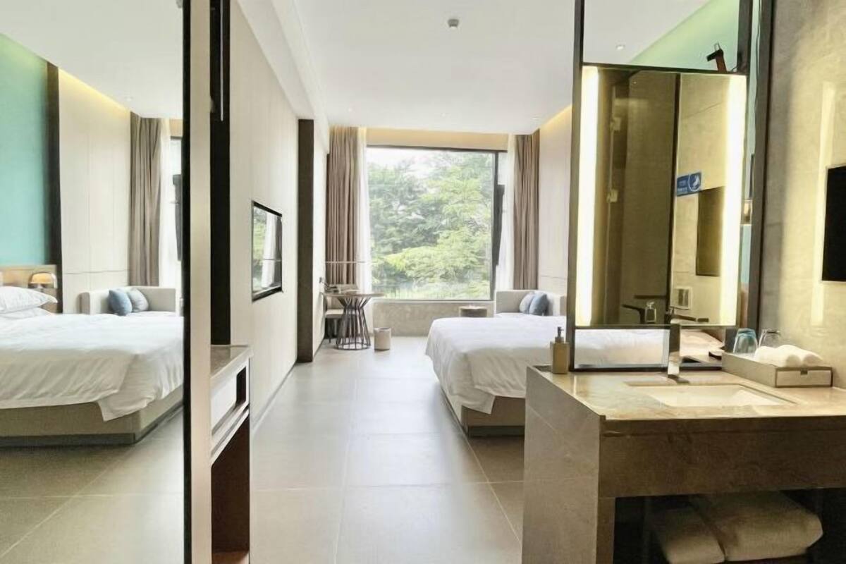 A hotel room with two beds, a large window, a bathroom with a mirror, and a small table with a chair.