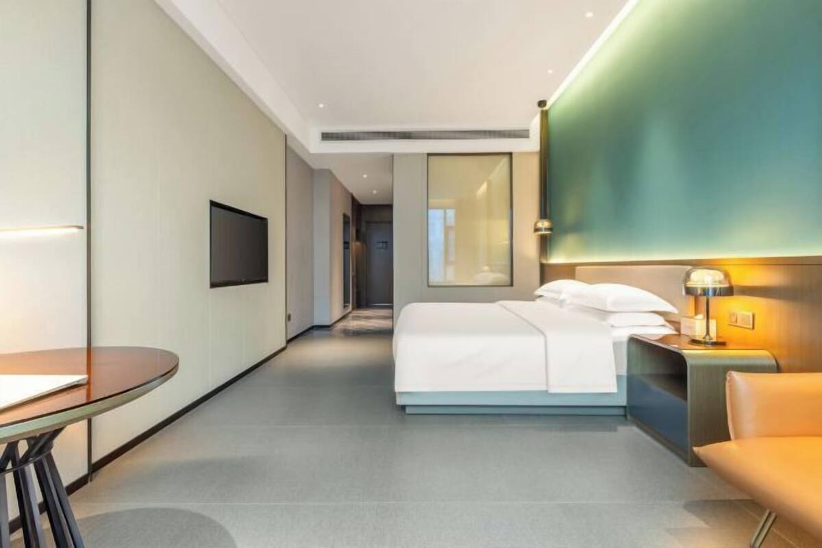 A modern hotel room with a bed, bedside table, lamp, and a TV mounted on the wall.
