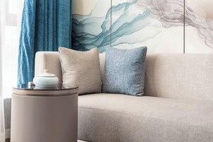 A modern living room with a beige sofa, two pillows, a round side table with a lamp, and a large abstract painting on the wall.
