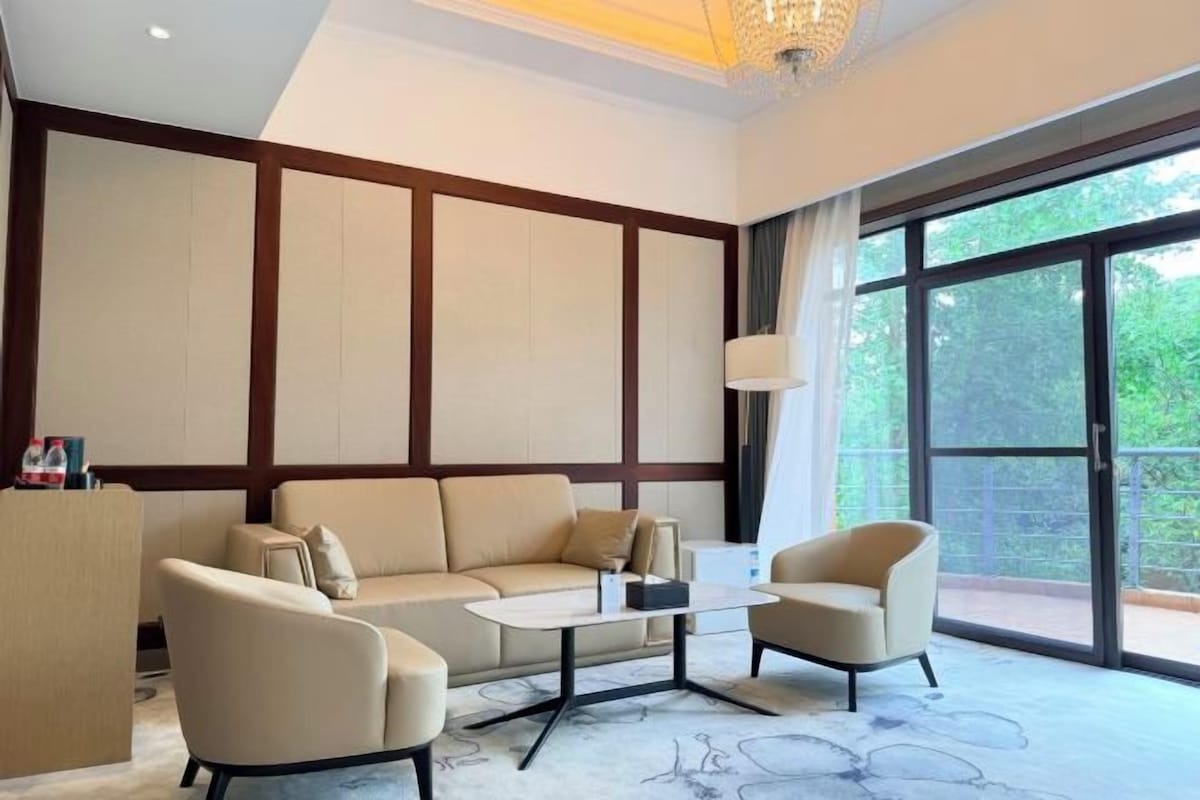 A modern hotel room with a sofa, armchairs, a coffee table, and a large window offering a view of greenery.