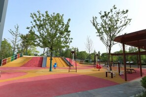 Un parque infantil con toboganes y columpios, rodeado de árboles y una zona de descanso cubierta.