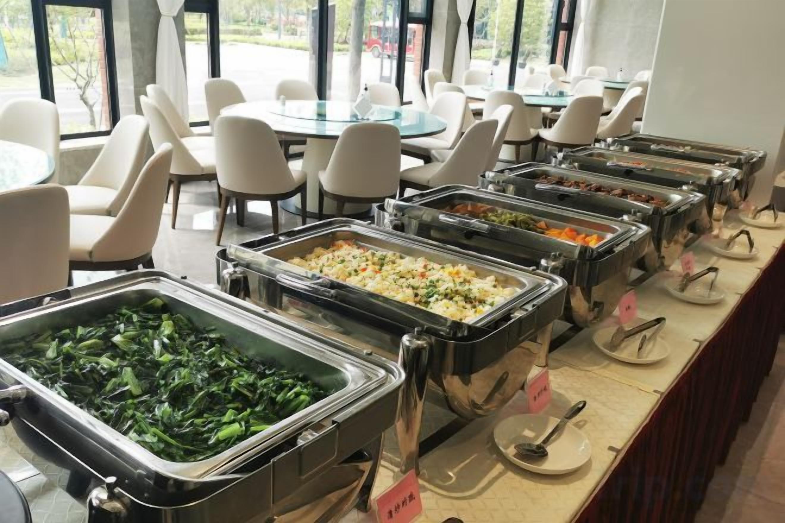 Montaje de bufé con diversas opciones de comida, incluyendo ensaladas y platos calientes, en un comedor con sillas y mesas.