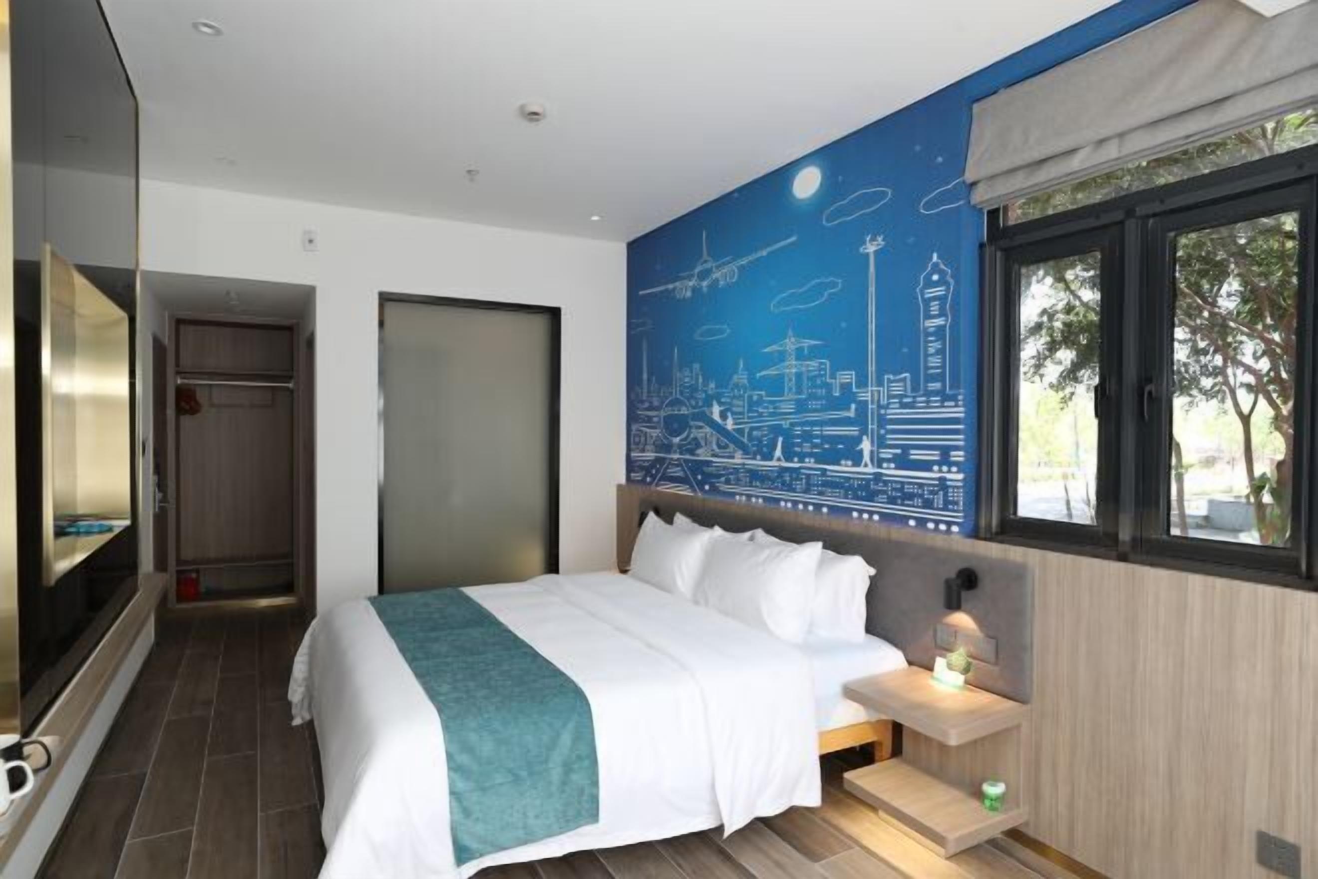 A modern hotel room with a large bed, a cityscape mural on the wall, a window with a view, and a small bedside table.