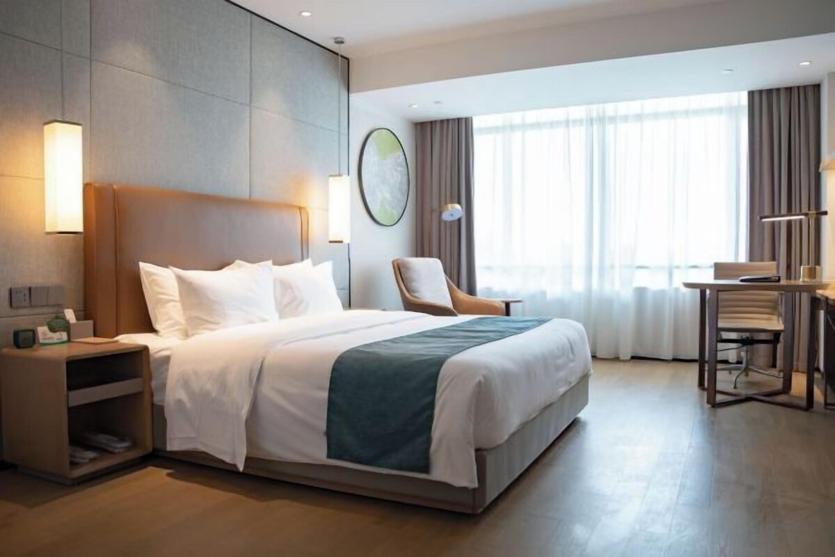 A hotel room with a large bed, bedside table, lamp, chair, and a view of the outdoors through the window.