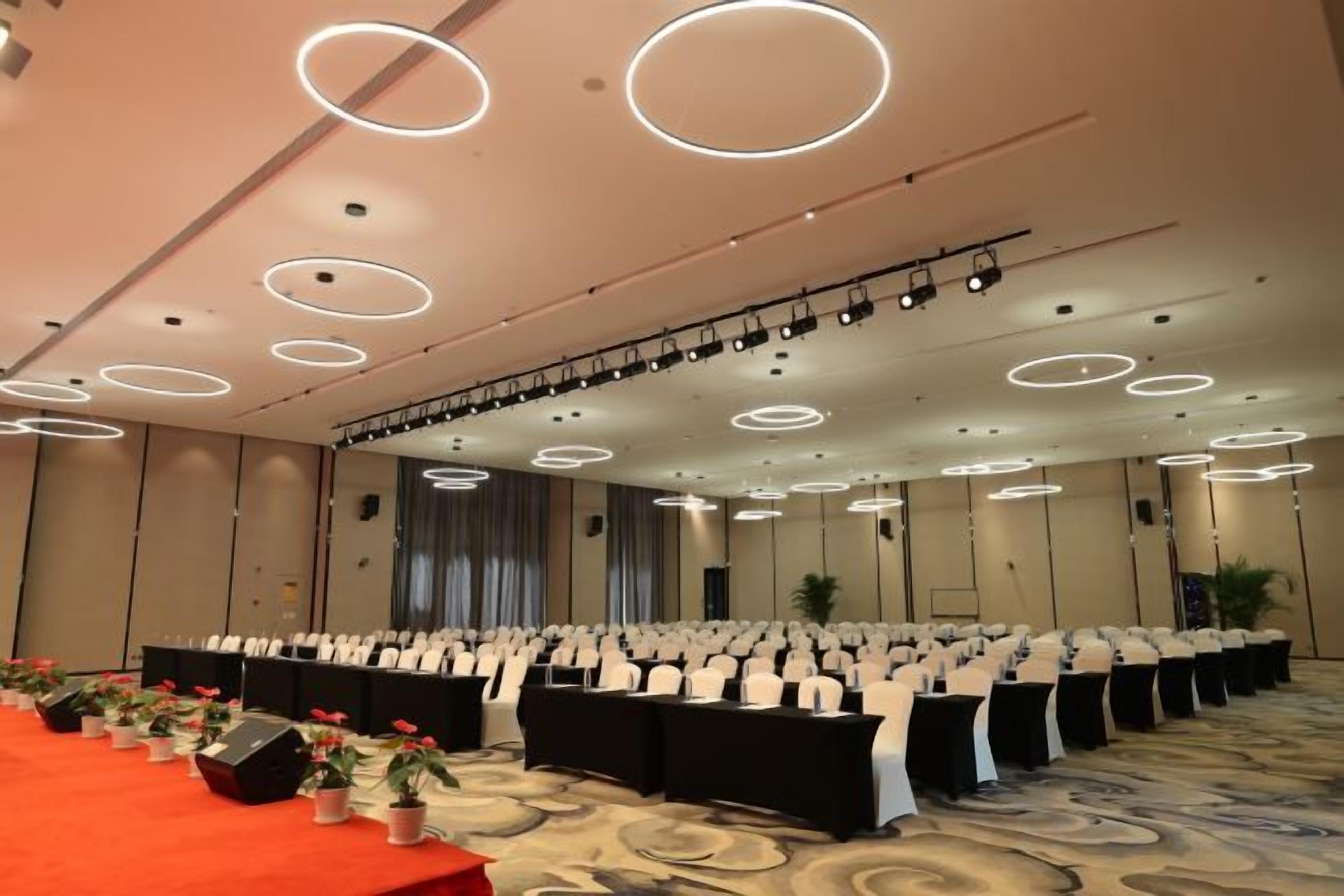 A conference room with rows of tables and chairs, a stage with lighting, and a carpeted floor.