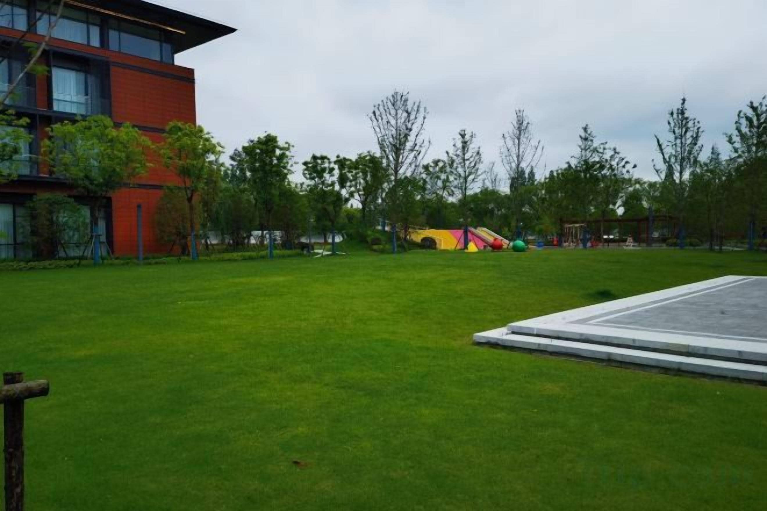 Un espacio con césped, un parque infantil, árboles y un edificio de fachada roja.