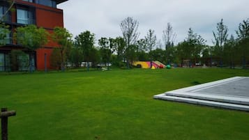 Un espacio con césped, un parque infantil, árboles y un edificio de fachada roja.