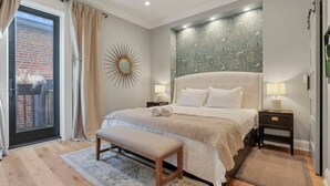Une chambre avec un grand lit, un banc, une table de chevet, une décoration murale représentant un soleil et une vue sur un bâtiment en briques, visible à travers une porte vitrée.