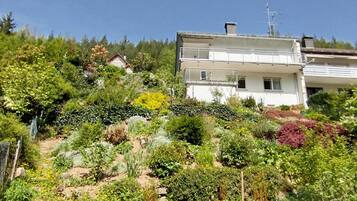Eine Wohngegend an einem Hang mit einem weißen, zweistöckigen Haus mit Balkonen, umgeben von vielfältiger Vegetation und einem klaren, blauen Himmel.