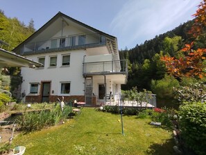 Een huis met twee verdiepingen, een balkon, een kleine tuin en uitzicht op een bebost heuvel.'