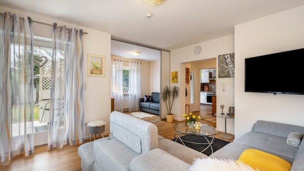 A modern living room with a sofa, a coffee table, and a TV. There is a dining area with a table and chairs visible in the background.
