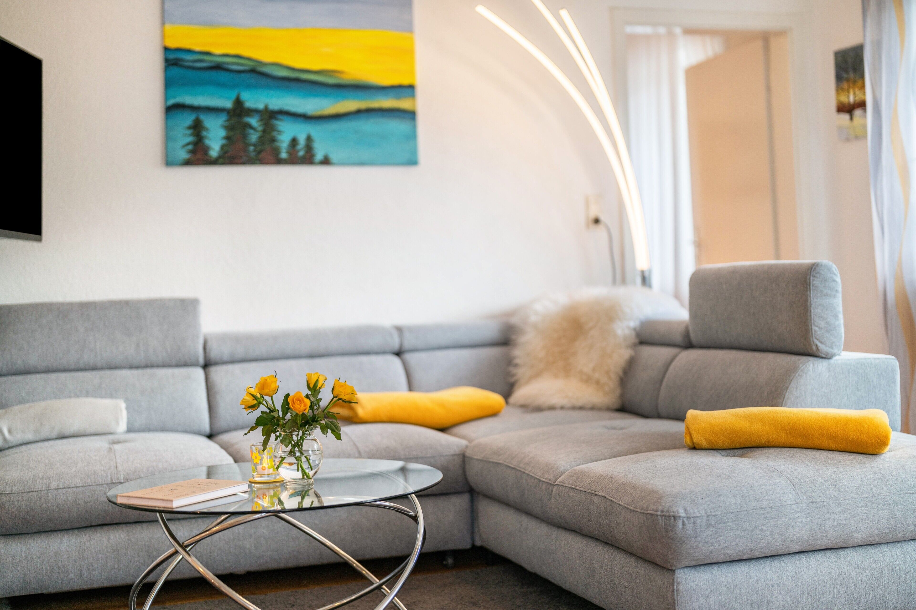 Een moderne woonkamer met een grijze bank, een glazen salontafel en een schilderij aan de muur.