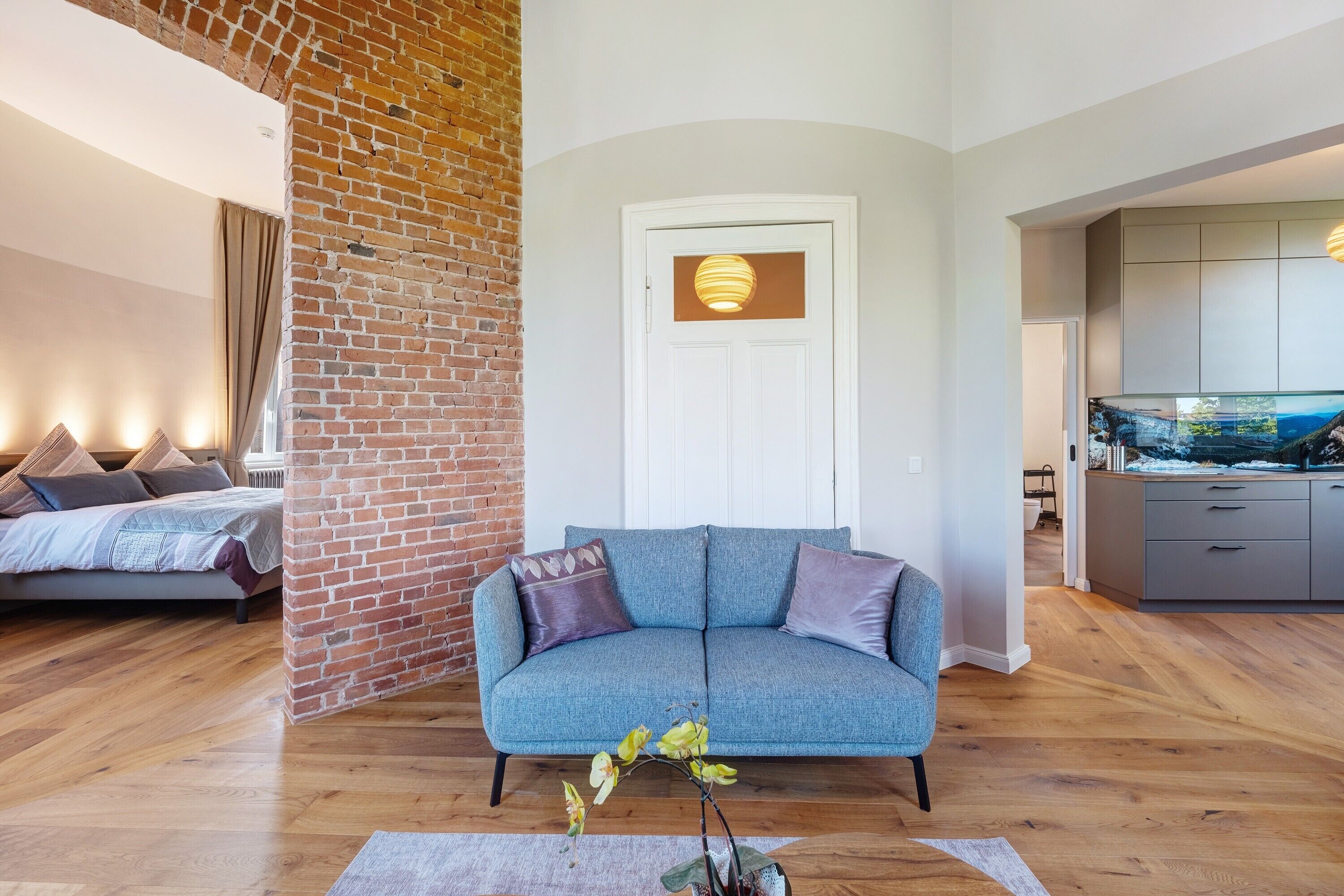 A modern living room with a blue sofa, a brick wall, a white door, and a kitchen area with a fish tank.