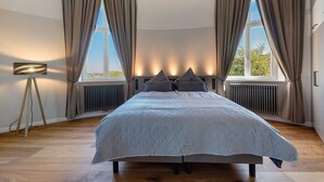 Une chambre avec un grand lit, deux fenêtres avec des rideaux, une lampe de sol et un radiateur.