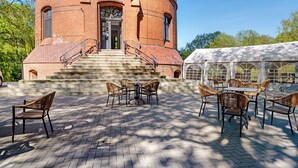 Un bâtiment en briques doté d’une tour circulaire, d’un espace extérieur aménagé avec des tables et des chaises, et d’une zone couverte sous une marquise blanche.