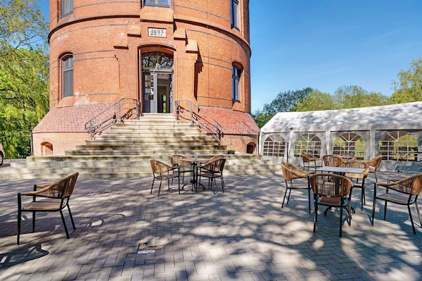A brick building with a circular tower, outdoor seating area with tables and chairs, and a covered area with a white canopy.