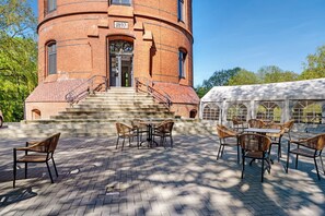 A brick building with a circular tower, outdoor seating area with tables and chairs, and a covered area with a white canopy.