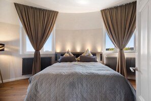 A bedroom with a large bed, two windows with curtains, a radiator, and a door.
