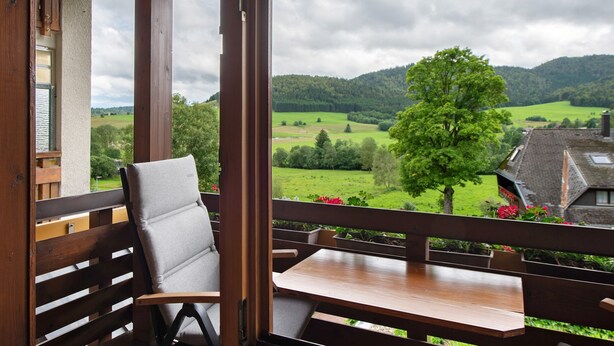 Een houten balkon met een tafel en stoel, met uitzicht op een groen landschap met bomen en een heuvel.