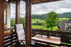 A wooden balcony with a table and a chair, overlooking a green landscape with trees and a hill.