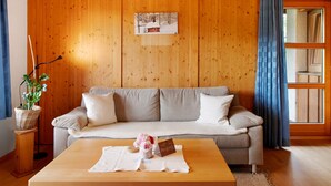 Een kamer met houten lambrisering, een bank, een tafel met een placemat, een mand met een plant en een ingelijst beeld aan de muur.