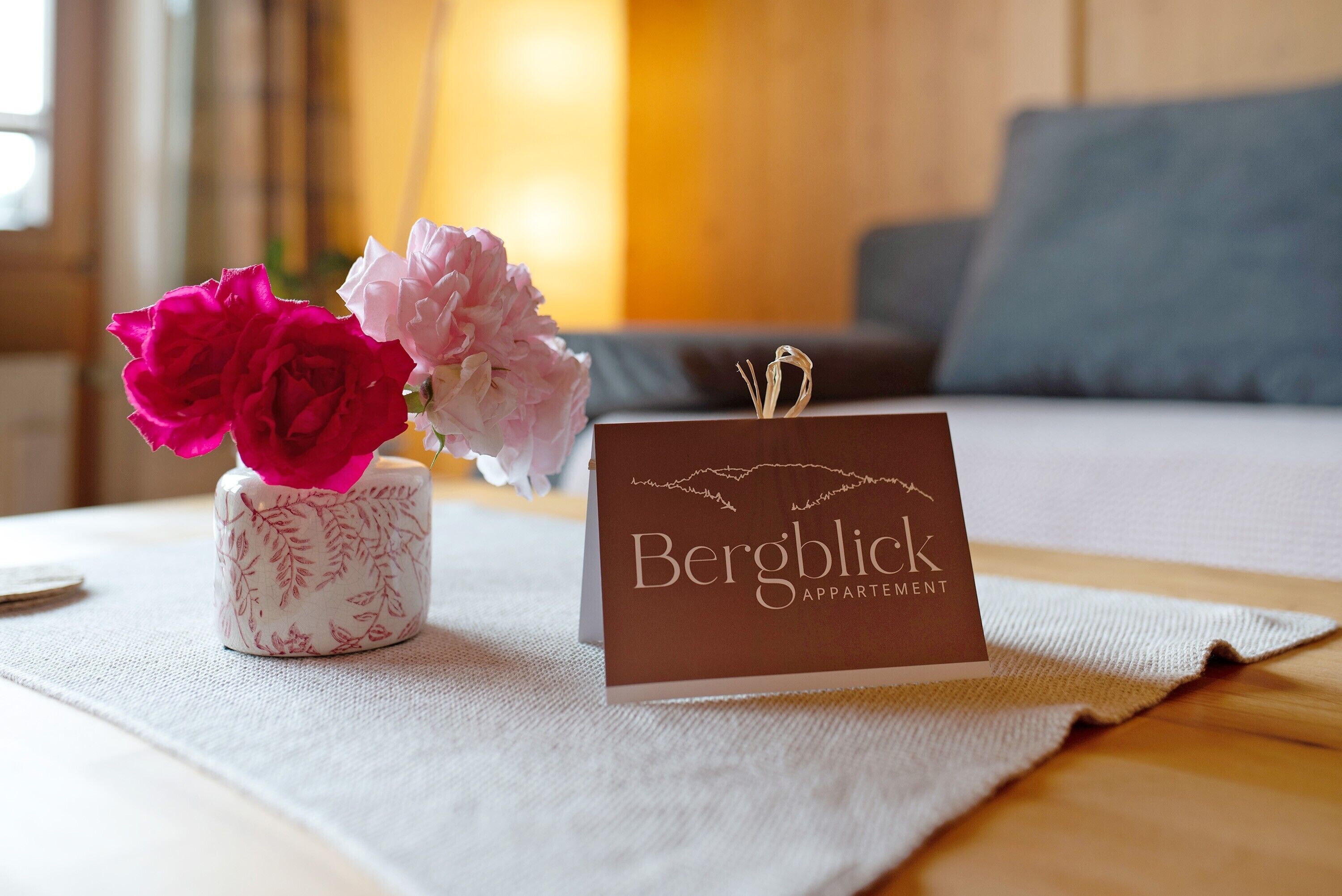Una pequeña mesa con una tarjeta de nombre que dice 'Bergblick Appartament' y un florero con flores rosas y blancas.