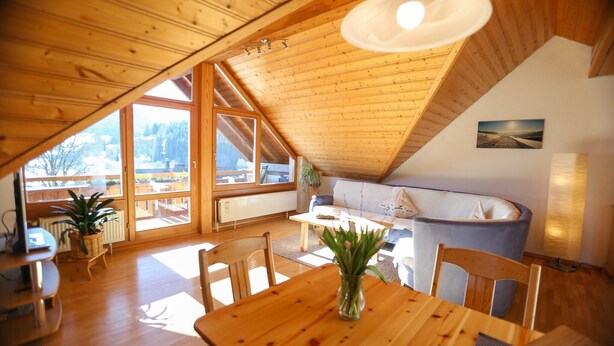A wooden interior with a dining table, chairs, a sofa, and a large window. - Apartment "Fichte" with Mountain View, Balcony, and Wi-Fi (Bernau im Schwarzwald)