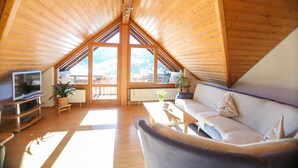 A cozy attic room with a wooden ceiling, a sofa, a TV, and a large window offering a view of the outdoors. - Apartment "Fichte" with Mountain View, Balcony, and Wi-Fi (Bernau im Schwarzwald)