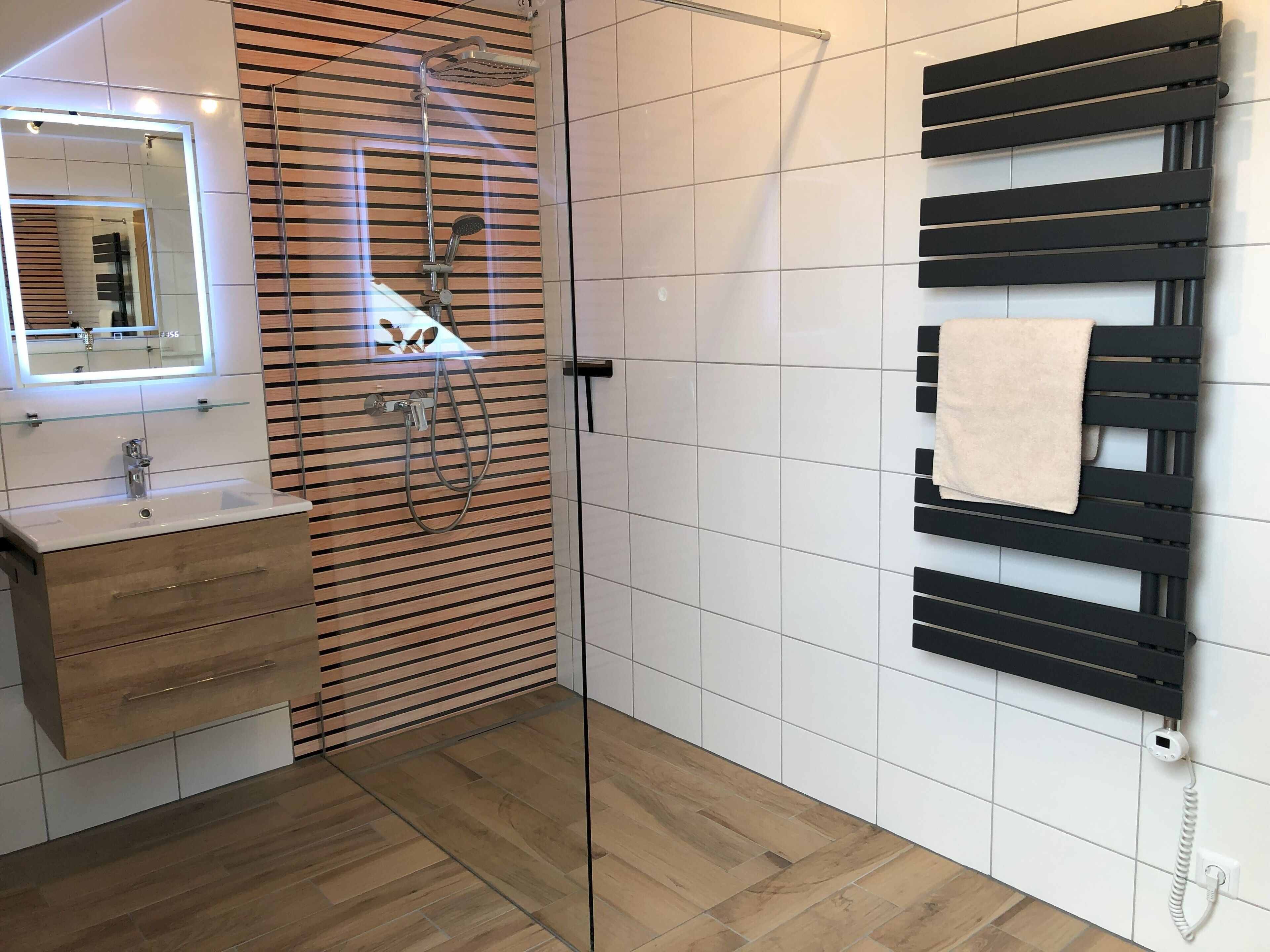 Et moderne badeværelse med brusekabine i glas, et træbord med vask, et spejl med LED-belysning og et håndklædetørrestativ.