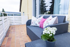 Un balcon avec un canapé en osier gris, des coussins blancs et une plante en pot.