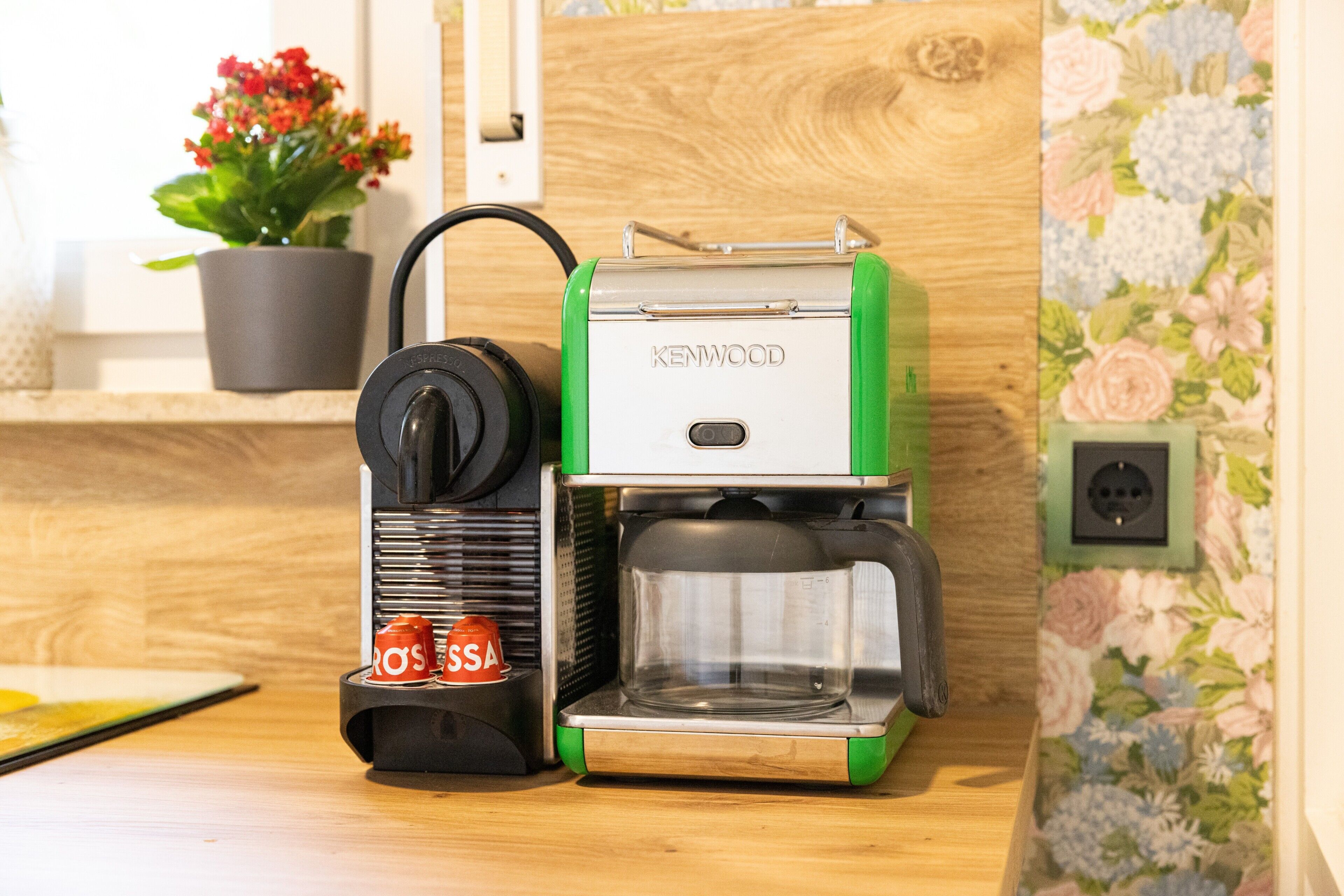Una cafetera con diseño verde y blanco, una jarra de café negra y una cápsula de café roja sobre una encimera de madera.
