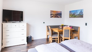 Una habitació amb un llit, un televisor sobre una dresser, una taula de dinar amb caires i dos quadres penjats a la paret.