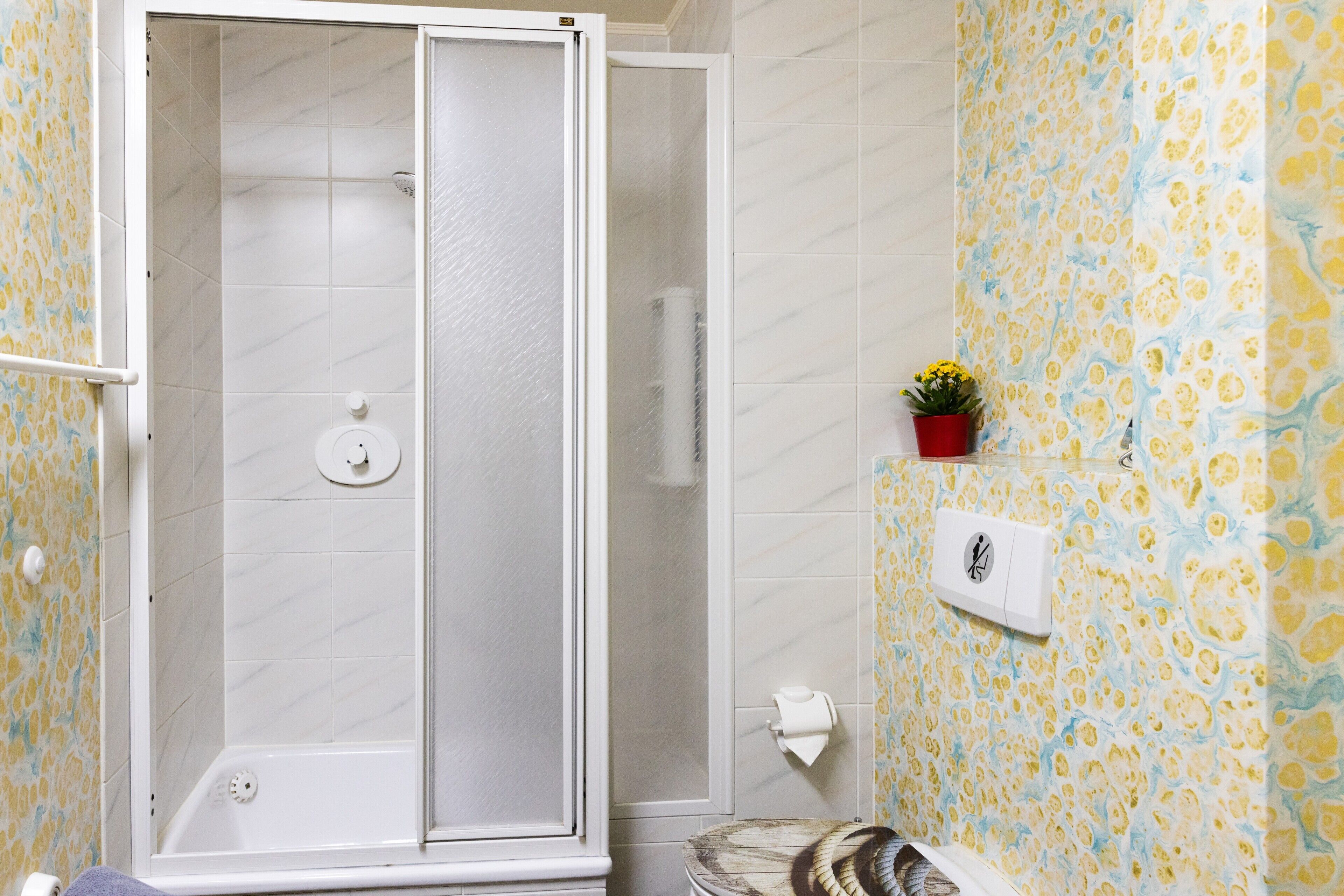 Une douche avec une porte en verre, des toilettes avec un abattant en bois et un mur recouvert de carreaux décoratifs.