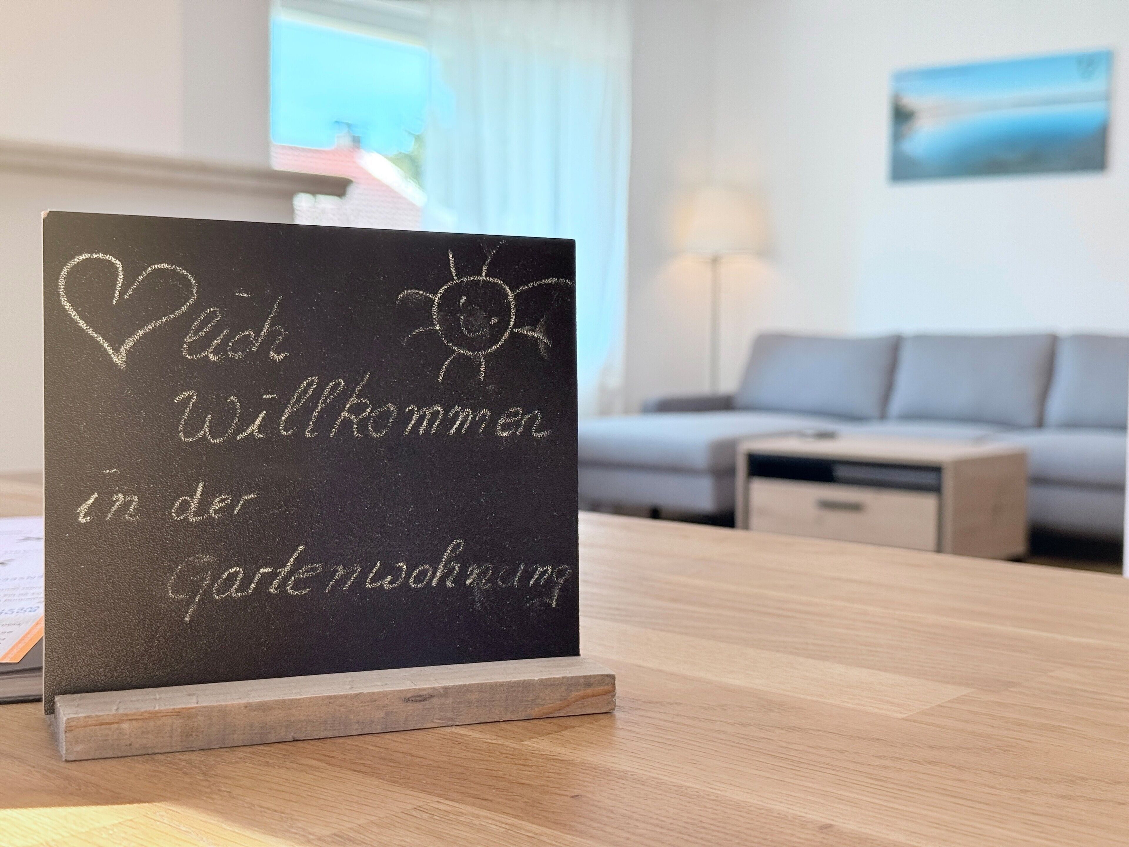 Una pissarra amb la inscripció 'Willkommen in der Gartenwohnung', col·locada sobre una taula de fusta en una habitació amb un sofà i una làmpada.