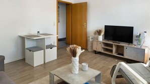 Una sala de estar moderna con televisor, una pequeña kitchenette y una puerta que da a otra habitación.