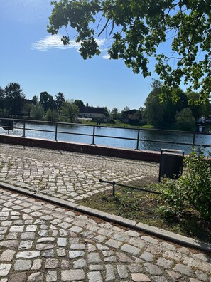 A cobblestone pathway beside a river with a metal railing, trees, and houses in the background.