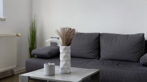 Una sala de estar moderna con un sofá gris, un jarrón blanco con plantas secas y una taza gris sobre una mesita.
