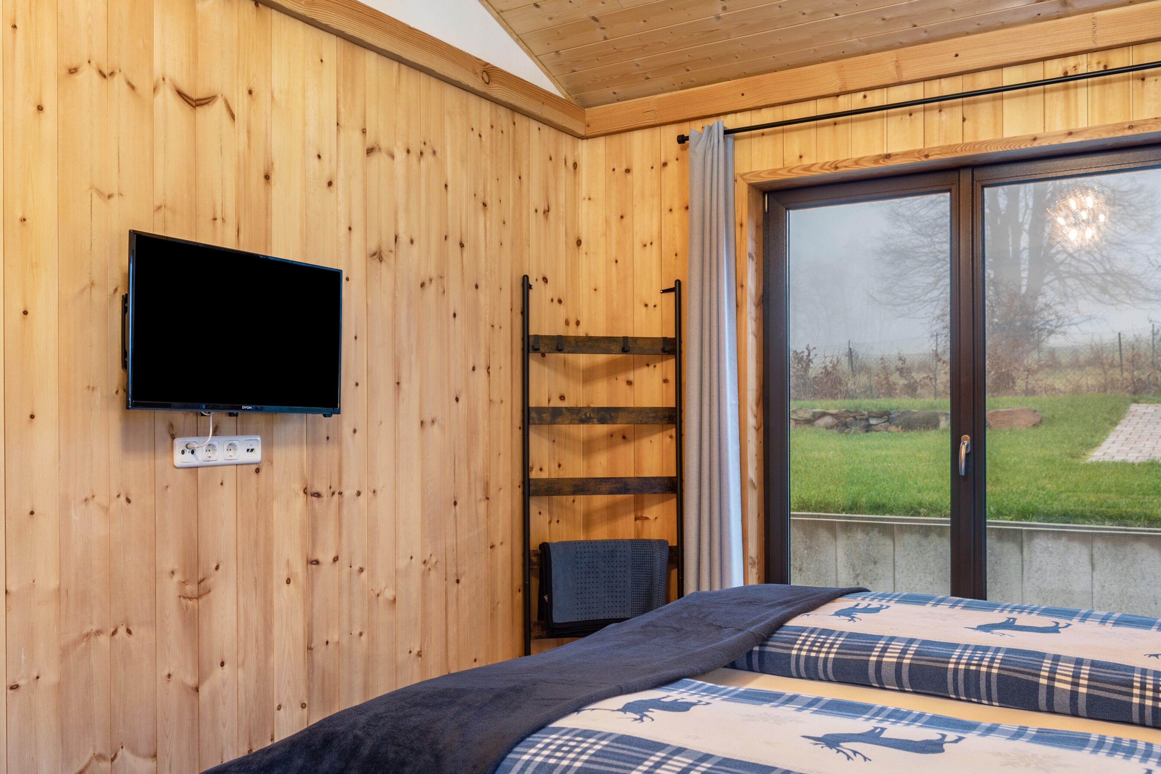 A wooden-paneled room with a bed, a television mounted on the wall, and a window with a view of the outdoors.