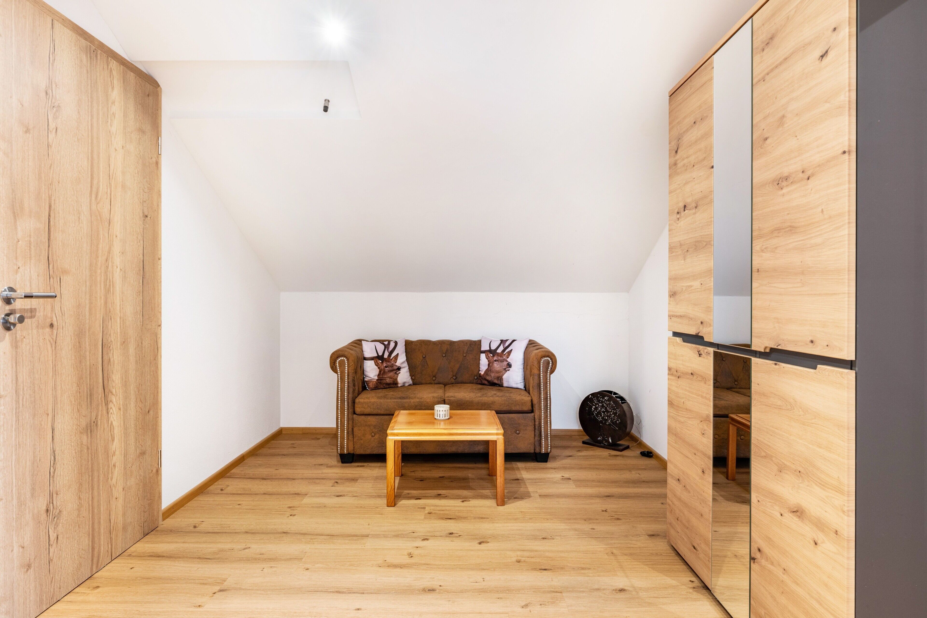 A room with a wooden floor, a sofa with decorative pillows, a small wooden table, and a large mirror.