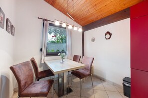 A dining area with a table, chairs, a window with curtains, a clock, and a trash bin.