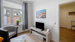 Un salon avec un téléviseur posé sur un meuble en bois, un fauteuil, une table avec des journaux, une fenêtre avec des rideaux et un tableau accroché au mur.