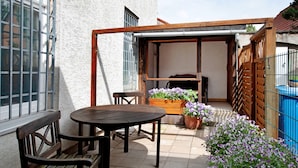 Une terrasse avec une table et des chaises en bois, un banc en bois, un pot de fleurs contenant des fleurs violettes et une clôture bleue.