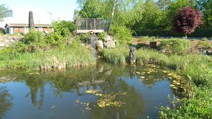 Un étang surmonté d’un pont en bois, entouré de verdure, avec un bâtiment visible au loin.