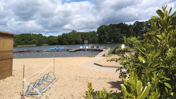 Une plage de sable avec un bateau bleu amarré à un quai, entourée de verdure et sous un ciel nuageux.