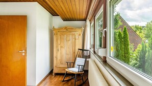 Une pièce avec une porte en bois, un fauteuil à bascule, une armoire en bois et une fenêtre donnant sur des arbres et un bâtiment.