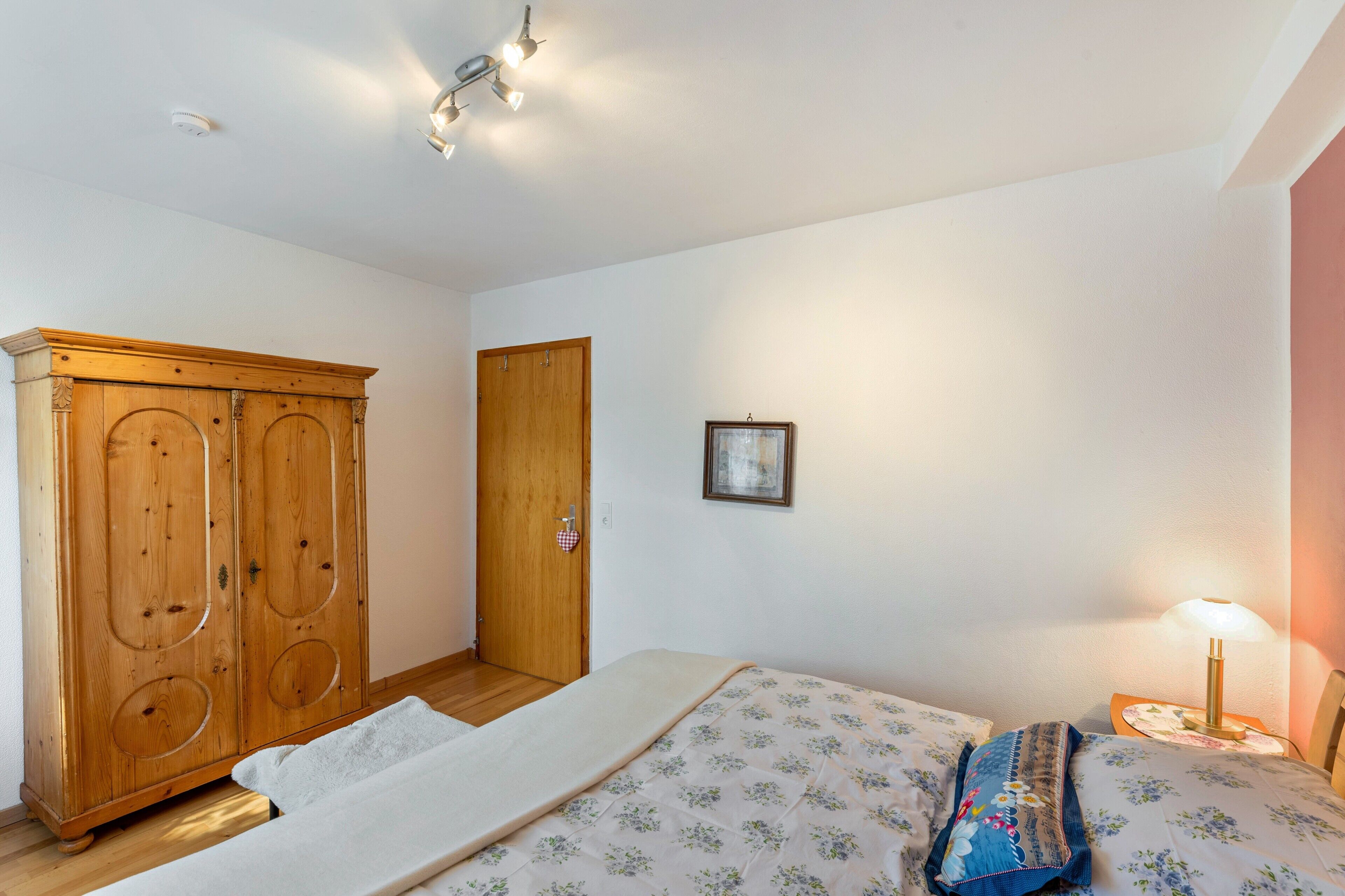 A bedroom with a bed, a wooden wardrobe, a door, a framed picture, and a bedside lamp.